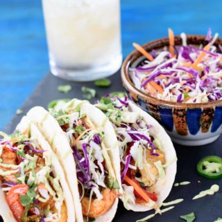 Three shrimp tacos on a slate platter with a margarita